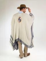 Poncho de hilo color crudo, con diseño de rayas azules y flecos en los bordes. Cuenta con un escudo bordado en la parte posterior.