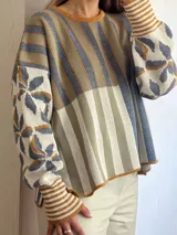 Sweater de tejido de punto con silueta oversize, cuello redondo y mangas largas abullonadas. Presenta un diseño patchwork con rayas verticales en tonos rojo, rosa y gris verdoso en el cuerpo, y un estampado floral en tonos rosa y rojo con detalles dorados en las mangas. Los puños y el cuello tienen un ribete a rayas horizontales.