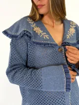 Saco tejido de punto color azul con diseño calado, cuello grande con bordados florales en tono crudo, volados decorativos y cierre frontal con un botón.