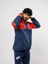 Campera de lluvia Umbro del Club Nacional de Football, con diseño de bloques de color en rojo y azul marino. Presenta capucha, cierre frontal, puños elásticos y detalles de piping blanco. Incluye logo de Umbro y escudo del club en el pecho.