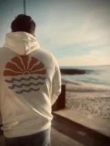 Hoodie de color crema con capucha y puños ajustados. Estampado trasero con un sol estilizado en color terracota y tres olas onduladas en color gris azulado.