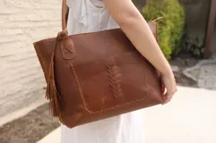 Cartera tipo tote de cuero vacuno color marrón, con diseño artesanal que incluye costuras decorativas en los laterales y en el bolsillo frontal. Cuenta con doble asa de hombro, cierre superior y un detalle de borla colgante en uno de los laterales.