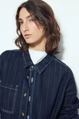 Sobrecamisa de denim azul oscuro con diseño de rayas verticales blancas, corte oversize, cuello clásico, cierre frontal con botones y bolsillos delanteros.