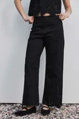 Pantalón negro de tiro alto y corte recto, confeccionado con detalles de bordados florales calados en la parte inferior de las piernas.