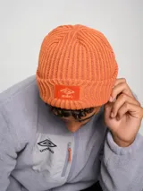Gorro tipo beanie de tejido acanalado en color azul, con parche rectangular rojo en el frente que presenta el logo de la marca Umbro bordado en blanco.