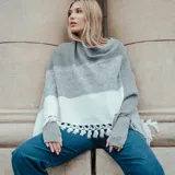 Poncho tejido de acrílico a rayas horizontales en tonos gris y blanco, con cuello amplio y mangas 3/4. Presenta detalle de flecos en el ruedo.