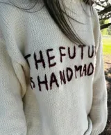 Sweater de lana merino virgen color crudo, con calce oversize y relajado. Presenta un bordado artesanal en el frente con la frase 'The Future Is Handmade' en color marrón.