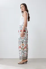 Pantalón de corte recto con estampado floral en tonos naranja, rojo, verde y marrón sobre fondo blanco. Presenta cintura elástica y ruedo con diseño geométrico.
