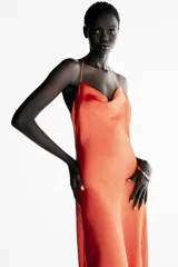 Vestido midi satinado de color naranja tostado, con escote fluido tipo cowl y tirantes finos cruzados en la espalda con detalle de aplicación metálica. Presenta un diseño largo con tajo lateral en la falda.
