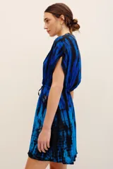 Vestido corto azul con estampado tie-dye en tonos negros y azules, escote en V cruzado, mangas cortas y cordón ajustable en la cintura. Presenta detalles de aplicaciones bordadas con lentejuelas en hombros y falda.