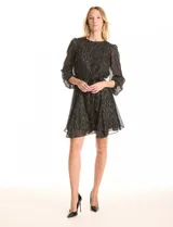 Vestido corto de gasa negra con estampado de rayas verticales doradas y plateadas. Tiene mangas largas semitransparentes con puños elastizados, cuello redondo, cintura con lazo ajustable y falda evasé con vuelo. La espalda presenta una abertura tipo lágrima con botón en la nuca.