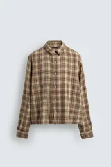 Camisa de corte cropped confeccionada en algodón, con estampado de cuadros en tonos beige, marrón y rojo. Presenta cuello solapa, cierre frontal de botones, manga larga con puño abotonado y detalles de roturas distribuidas por toda la prenda con un bajo irregular.
