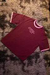 Remera oversize color bordó con cuello y puños de rib de color rosado. Estampado frontal en rosado con el texto "GUARDIÁN DE LA FUERZA" y otros símbolos.