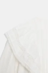 Camisa blanca de cuello redondo y manga larga, con detalle de volantes frontales y entredoses de encaje. Presenta un cierre en la espalda con abertura y botón.