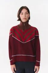 Buzo de tejido de punto en color azul marino, con cuello alto y cierre frontal parcial. Presenta un diseño con franjas en color marrón y detalles de líneas en rojo en el pecho, hombros y ruedo. Tiene corte holgado y mangas largas con puños acanalados.