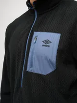 Buzo de entrenamiento Umbro con medio cierre frontal, cuello alto y bolsillo en el pecho de nylon con cierre. Presenta una textura sutil en el tejido y logo de la marca estampado en el bolsillo.