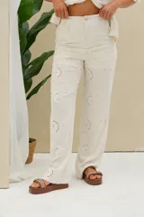 Pantalón largo color beige de corte recto, confeccionado con tejido bordado estilo broderie con motivos florales y perforaciones circulares. Cuenta con cintura alta y cierre frontal con botón y cierre.
