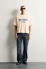 Camiseta relaxed fit de color beige, con cuello redondo, mangas cortas y estampado frontal en tono azul con texto gráfico.