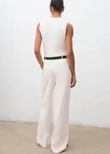 Mono largo de diseño recto y estilo minimalista, color blanco. Presenta cuello perkins, sin mangas con tirantes anchos, detalle de pinzas delanteras y cierre de cremallera en la parte posterior. Incluye bolsillos laterales y un cinturón negro con hebilla metálica.