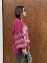 Buzo de tejido de punto con textura peluda, de corte holgado y mangas amplias. Presenta un diseño geométrico con una flor grande estampada en el centro, combinando tonos fucsia, rosa y marrón en un patrón de bloques y rayas.