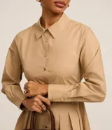 Vestido corto color beige, estilo chemise, confeccionado en tricolina. Tiene cuello inglés, mangas largas con puños abotonados, abotonadura frontal y falda con pliegues.