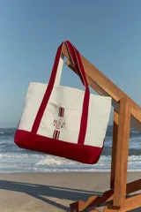 Bolso tipo totebag de lona color crudo con base y asas en color rojo. Estampado frontal con el texto "The Beach Club" en rojo.