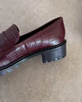 Mocasines negros de estilo clásico con textura de cuero grabado tipo cocodrilo. Presentan una banda frontal característica y suela plana con borde definido.