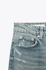 Jeans de tiro alto con diseño mom fit, confeccionados en denim de lavado medio con efecto desgastado y roturas localizadas. Presentan cinco bolsillos, trabillas para cinturón y cierre frontal con cremallera y botón metálico.
