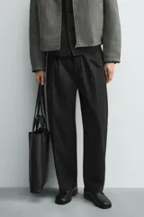 Pantalón negro de corte relaxed fit y estilo wide leg, con pliegues frontales dobles, bolsillos delanteros y detalle de bolsillos traseros de vivo. Cierre frontal cruzado con cremallera y gancho metálico oculto.