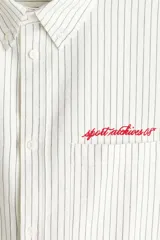 Camisa de corte holgado en mezcla de algodón Oxford, con diseño a rayas, cuello americano y cierre frontal de botones. Cuenta con un bolsillo superior en el pecho con texto bordado en rojo, mangas largas con puños abotonados y bajo recto.