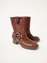 Botas de caña media confeccionadas en cuero texturado color marrón. Presentan un diseño robusto con múltiples hebillas metálicas ajustables, apliques circulares laterales y punta cuadrada. Cuentan con un taco cuadrado de 5 cm de altura.