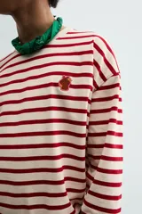 Camiseta de manga larga con diseño de rayas horizontales en color rojo y crema. Presenta cuello redondo con ribete a contraste y un pequeño parche bordado en el pecho.