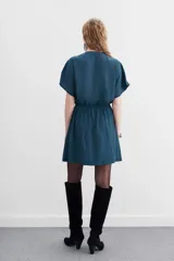 Vestido mini de escote cruzado en color azul petróleo, con mangas cortas y cintura elastizada con lazo ajustable. Presenta detalles de cadenitas metálicas decorativas en los hombros y en la terminación del lazo.