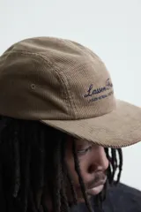 Gorra de cinco paneles confeccionada en pana de algodón, con visor plano y ajuste posterior. Presenta un bordado frontal con el texto 'Lassen Field'.