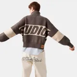 Campera de tejido de punto marrón con cierre frontal y cuello tipo polo. Presenta un diseño de bloques de color con franjas beige en el pecho y los brazos, y la palabra "EME" estampada en grande en el frente.