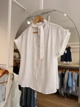 Blusa blanca de rayón y poliéster, con escote en V, detalle de lazo en el cuello, botonadura frontal y mangas cortas con vuelta.
