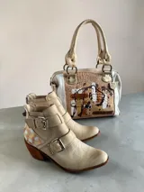 Bota corta de cuero natural color beige, con taco de madera, puntera afinada y cierre trasero. Cuenta con tiras con hebillas metálicas y detalles pintados a mano en la parte trasera.