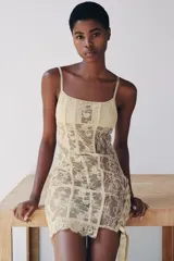 Vestido corto semitransparente tipo corsetero color beige, confeccionado con encaje y detalles de varillas interiores que estilizan la figura. Presenta escote redondo, tirantes finos ajustables y bajo asimétrico con una lazada lateral de satén.