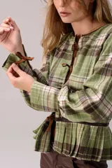 Blusa de tartán con diseño versátil, confeccionada en tela escocesa con detalles en cuero de oveja. Presenta cuello alto con forro de piel sintética, mangas largas con puños de cuero y piel, y un ruedo tableado con ajuste de cinturón de cuero en la cintura.