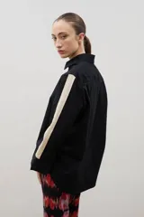 Camisa de corte holgado y largo medio, color negro con detalle de franja lateral en color crema a lo largo de las mangas. Presenta cuello clásico, cierre frontal con botones y tajos laterales en el ruedo.