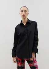 Camisa de corte holgado y largo medio, color negro con detalle de franja lateral en color crema a lo largo de las mangas. Presenta cuello clásico, cierre frontal con botones y tajos laterales en el ruedo.