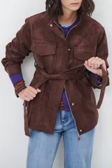 Chaqueta de gamuza en tono marrón oscuro, con cuello alto, cierre frontal con cremallera y botones a presión, bolsillos frontales con solapa y cinturón ajustable en la cintura.
