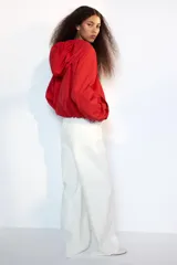 Chaqueta corta de nylon color beige, con cuello subido, capucha y cierre frontal con cremallera. Presenta mangas largas con puños elásticos, bolsillos laterales y detalles de cordones ajustables con lazadas en el pecho y el bajo.