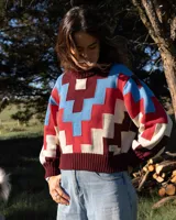 Sweater de cuello alto confeccionado en tejido de punto con diseño geométrico en bloques de colores bordó, rojo, celeste y beige. Presenta puños y ruedo acanalados y un pequeño parche decorativo en el frente.