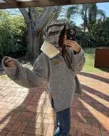 Buzo canguro oversize de lana gris, con cuello forrado en corderito color crema y capucha tejida con patrón multicolor en tonos tierra y blanco. Presenta detalles de cuero marrón en el cierre frontal y en los laterales.