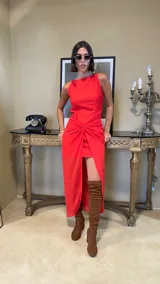 Vestido largo rojo con diseño asimétrico, escote redondo y detalle de nudo drapeado en la cintura que crea una abertura frontal, dejando ver una falda corta interior.