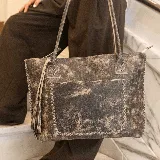 Cartera tipo tote de cuero marrón con costuras artesanales visibles, bolsillo frontal con el logo grabado, asas de hombro y detalles de flecos colgantes.