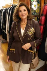 Blazer de gamuza color tostado con diseño artesanal. Presenta flecos en las solapas y bolsillos, bordados florales en los hombros con detalles de perlas colgantes y aplicaciones decorativas en los bolsillos.