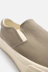 Zapatilla tipo slip-on de lona color taupe, con elástico lateral para facilitar el calce y suela de goma gruesa color crema.