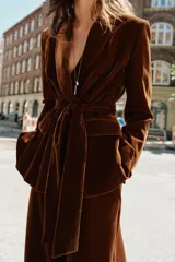 Blazer de terciopelo color marrón, con cuello solapa, manga larga y cierre frontal cruzado con botones. Incluye bolsillos con solapa en el delantero y un cinturón del mismo tejido para ajustar a la cintura.